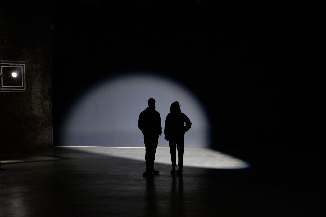 Two people are silhouetted in a dark room with a spotlight hitting part of a black wall behind them and the concrete floor.